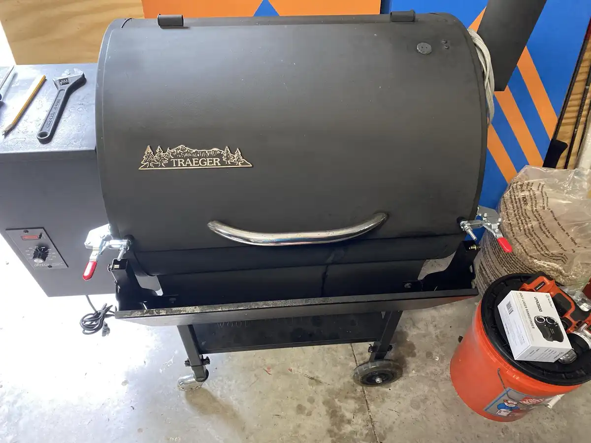 A black Traeger wood pellet grill sits on a wheeled cart in a garage, with a bag of pellets and a box of a golf laser…
