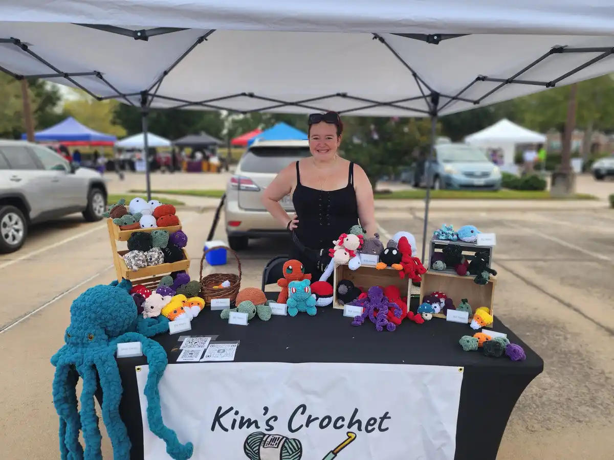 A smiling woman stands behind a table at an outdoor market under a white canopy, surrounded by numerous colorful crocheted…