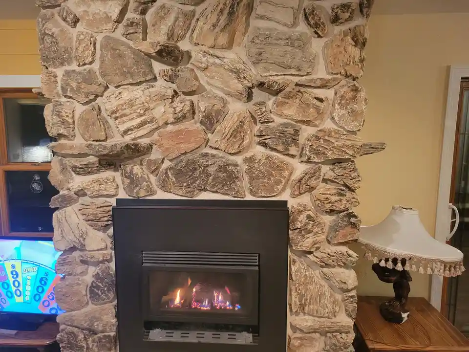 A cozy room features a stone fireplace with a lit gas fire, next to a lamp and a TV showing a colorful game show wheel. The…