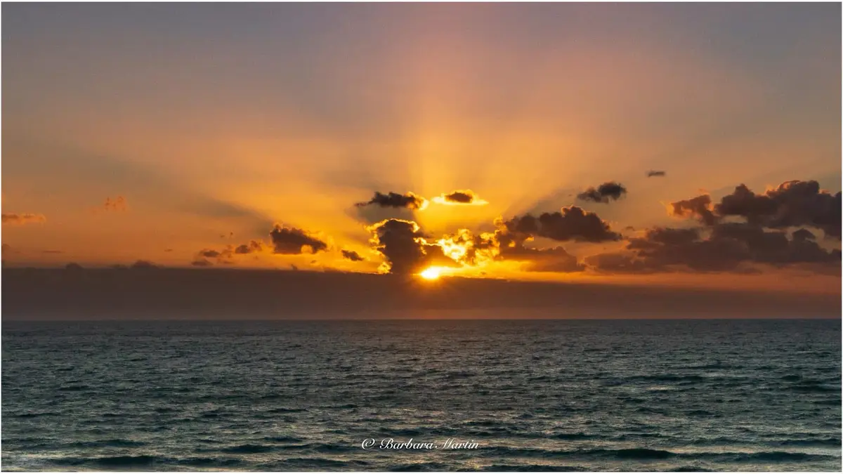 A serene photograph of a sunrise over a calm ocean, with the sun partially obscured by clouds and golden rays bursting…