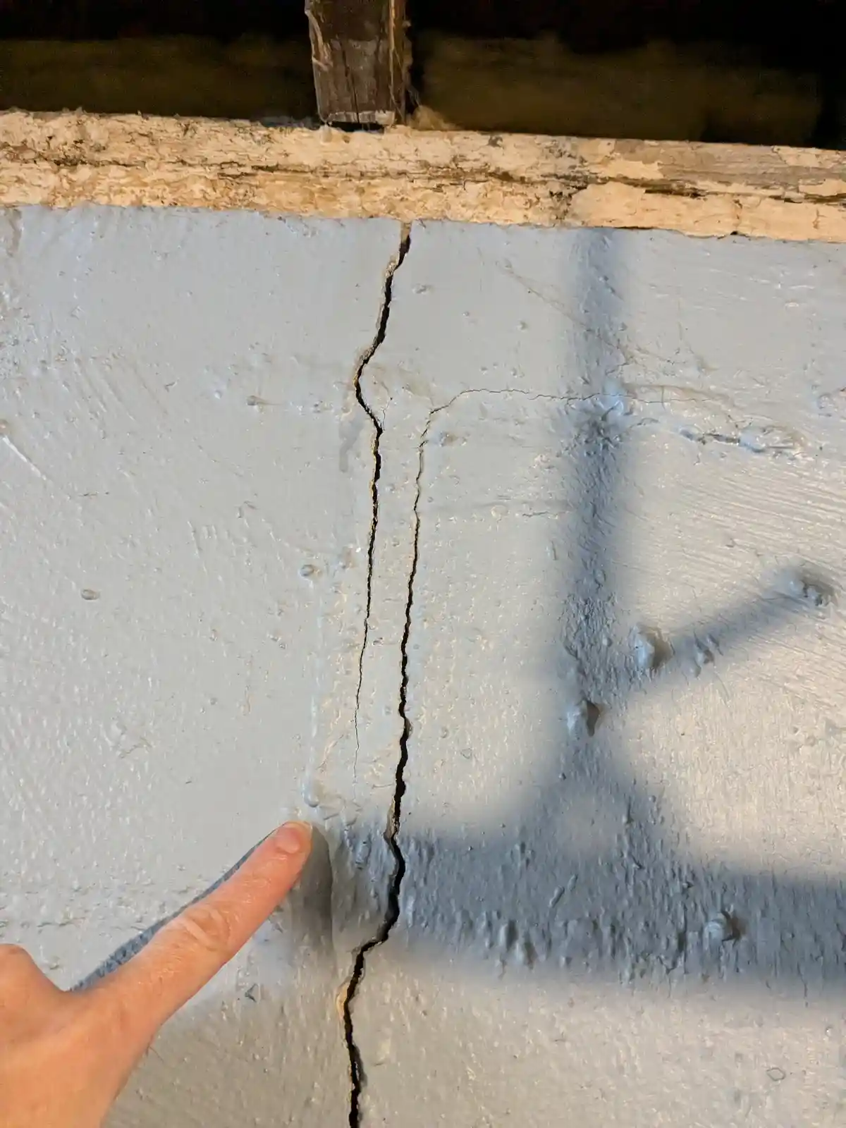 A close-up photo shows a finger pointing at a prominent vertical crack running through a light gray painted wall, with a…
