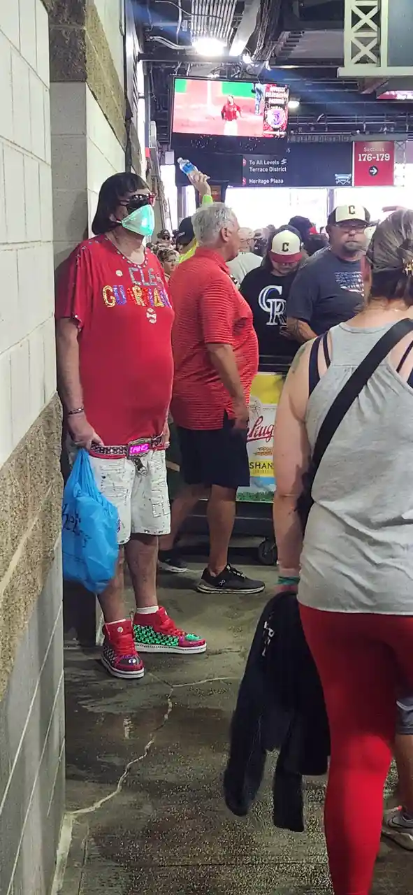 A person in a Cleveland Guardians shirt, wearing a mask, sunglasses, and brightly colored sneakers, stands in a crowded…