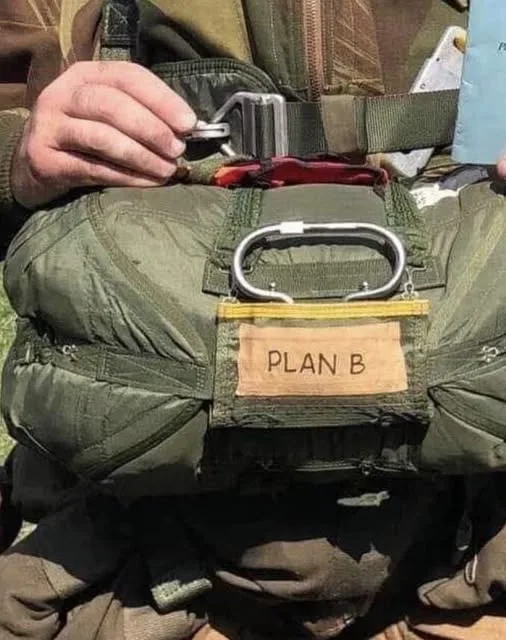 A close-up photo shows a person in military-style gear holding a folded parachute. A tan label stitched onto the parachute…