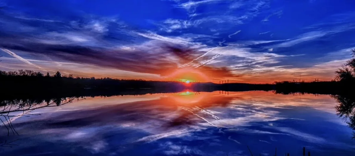 A vibrant, panoramic sunset over a calm lake, with the sky painted in deep blues and fiery oranges, mirrored perfectly in…