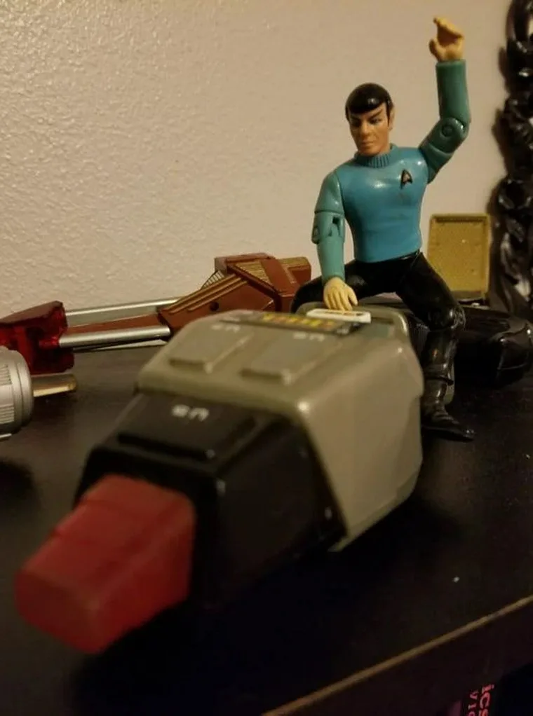 A plastic toy figure of Spock from Star Trek, wearing his blue uniform and black boots, is posed sitting on a gray and black…