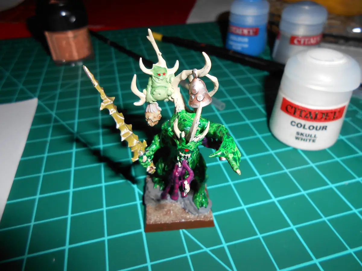 A green-painted fantasy miniature of a horned demon-like creature with a smaller green figure on its head, both with red…