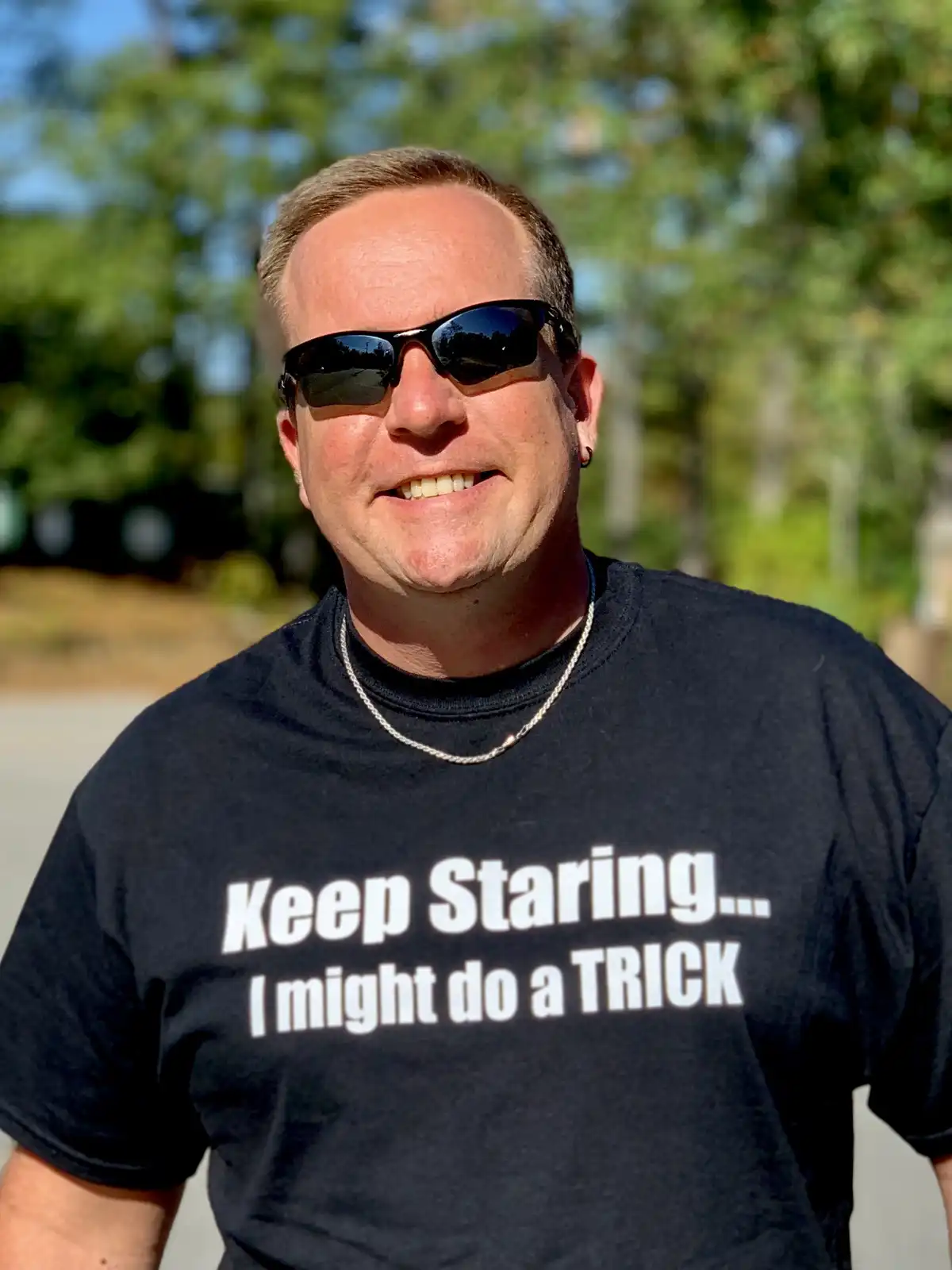 A man wearing sunglasses and a black t-shirt with white text that reads 'Keep Staring... I might do a TRICK' smiles at the…