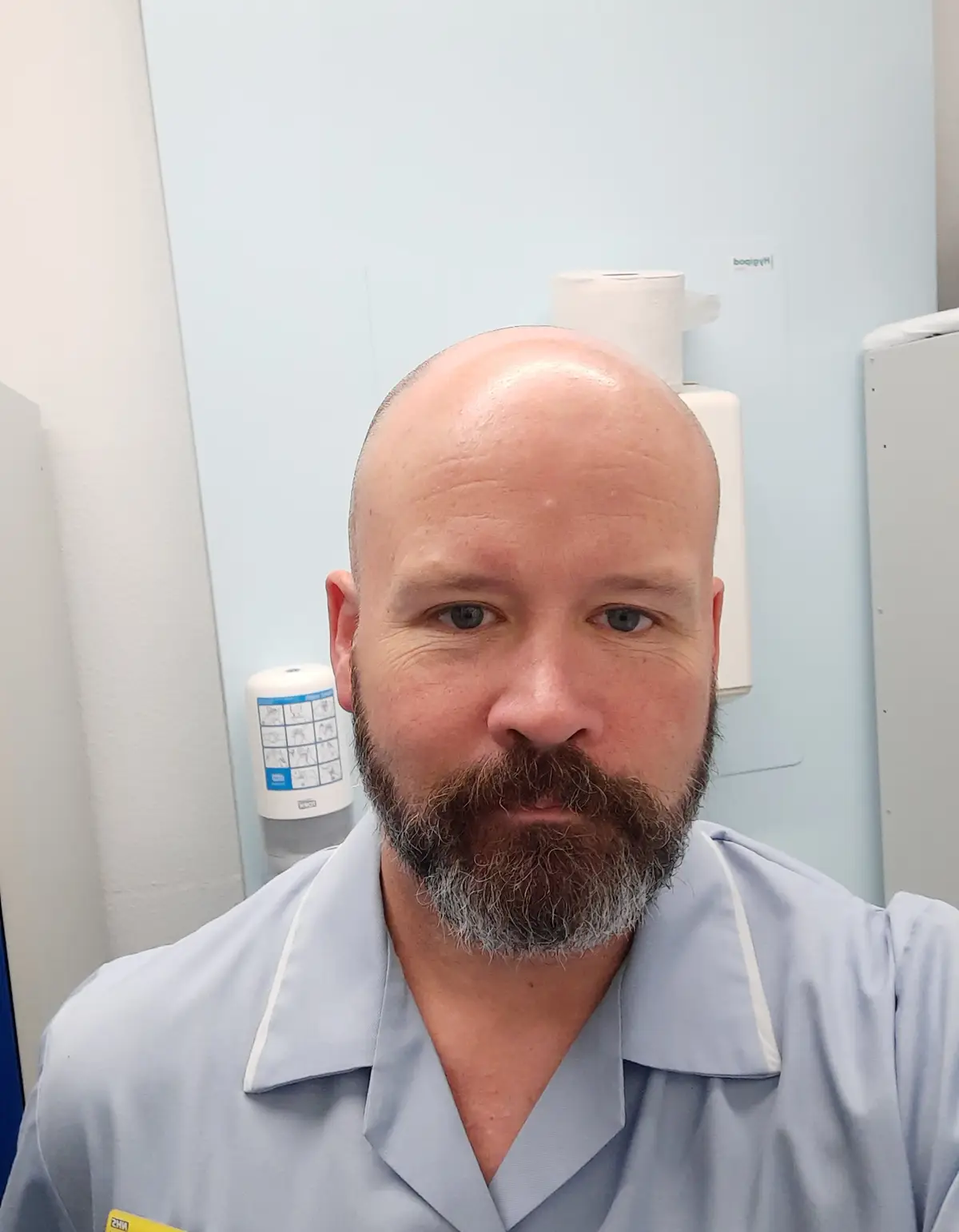 A bald man with a full beard, wearing light blue medical scrubs, stares directly at the camera with a serious expression…