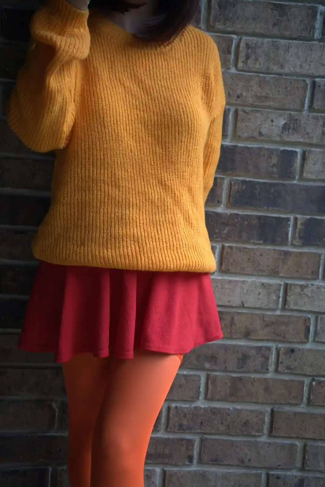 A person wearing a mustard yellow ribbed sweater, a red pleated skirt, and orange tights stands against a brick wall. The…