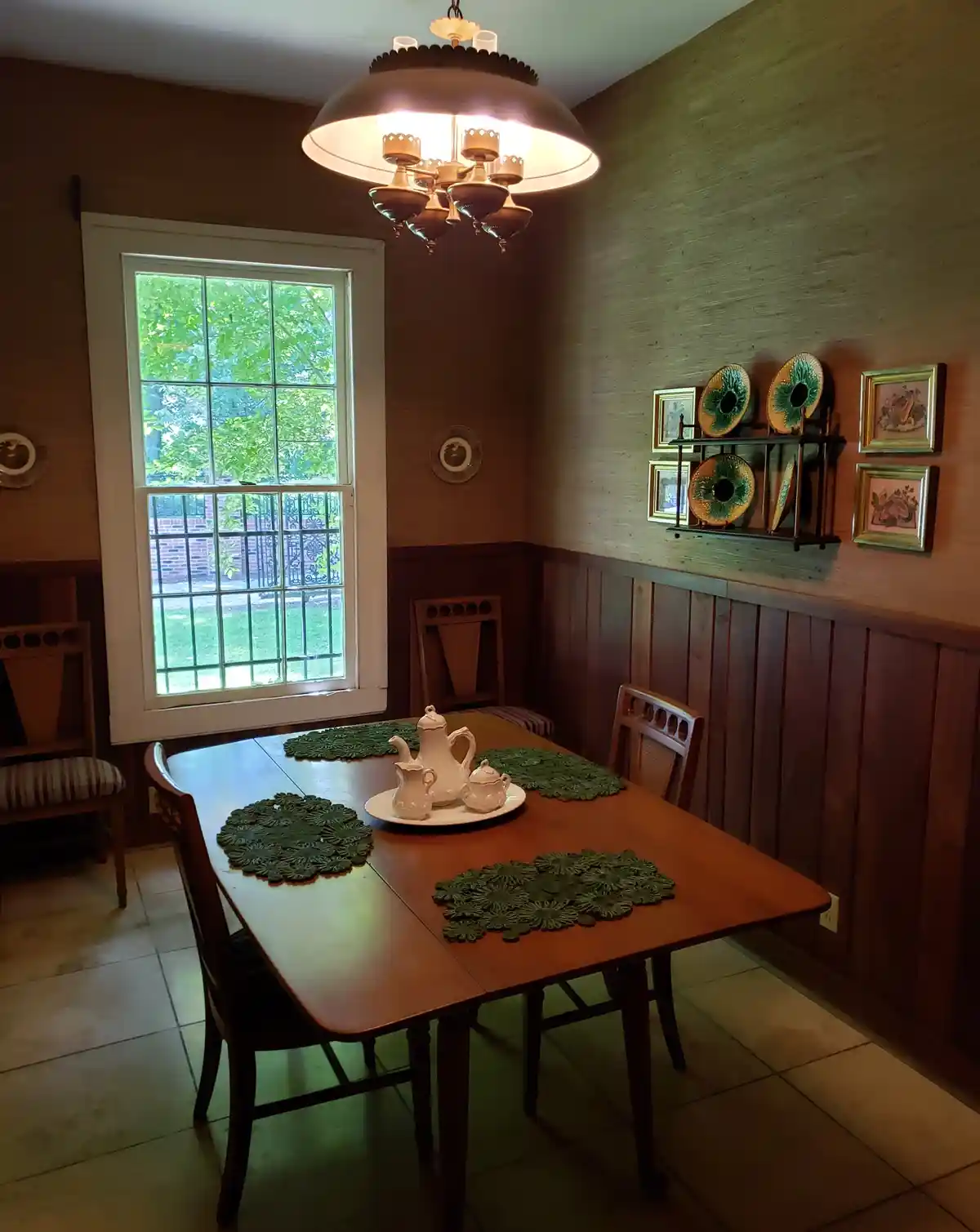 A warmly lit, vintage-style dining room with a wooden table set for tea, featuring green floral placemats and a white…