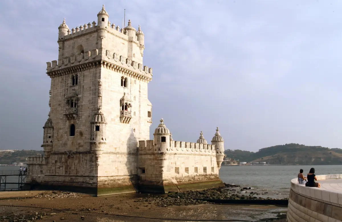 A wide shot of the historic Belem Tower in Lisbon, Portugal, standing by the water with a few tourists nearby. The overlaid…