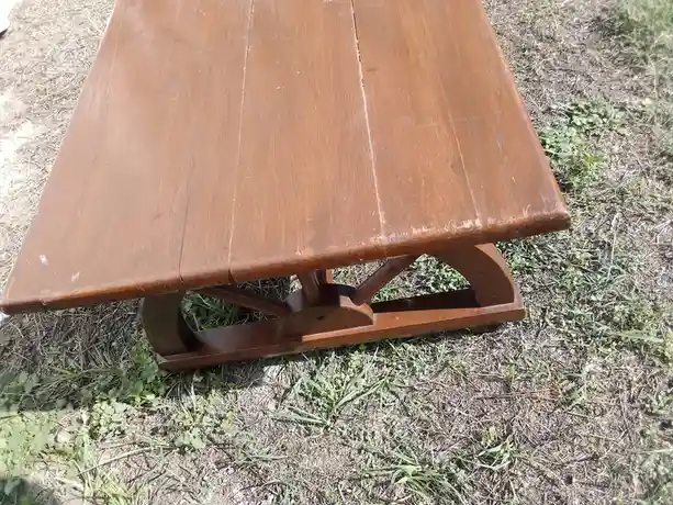 A close-up photo shows a brown wooden table with a distinctive curved base, resting on dry grass and sparse green weeds. The…