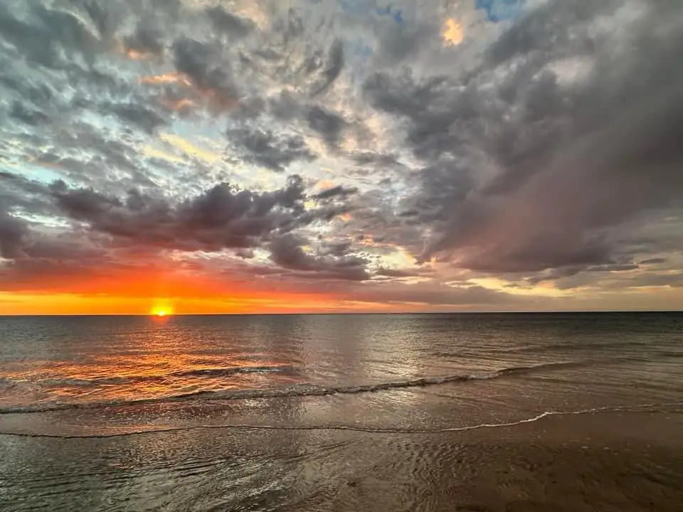 A serene beach sunset with orange and purple clouds, the sun low on the horizon over calm water, with gentle waves lapping…