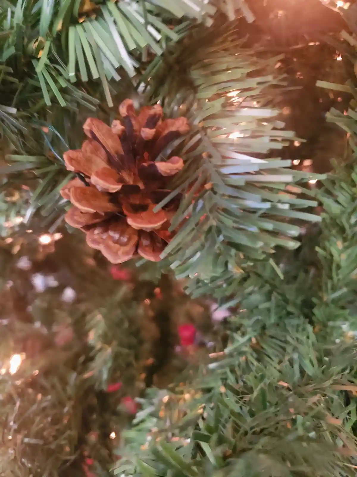 A close-up photo shows a brown pinecone nestled among the green, artificial needles of a decorated christmas tree, with warm…