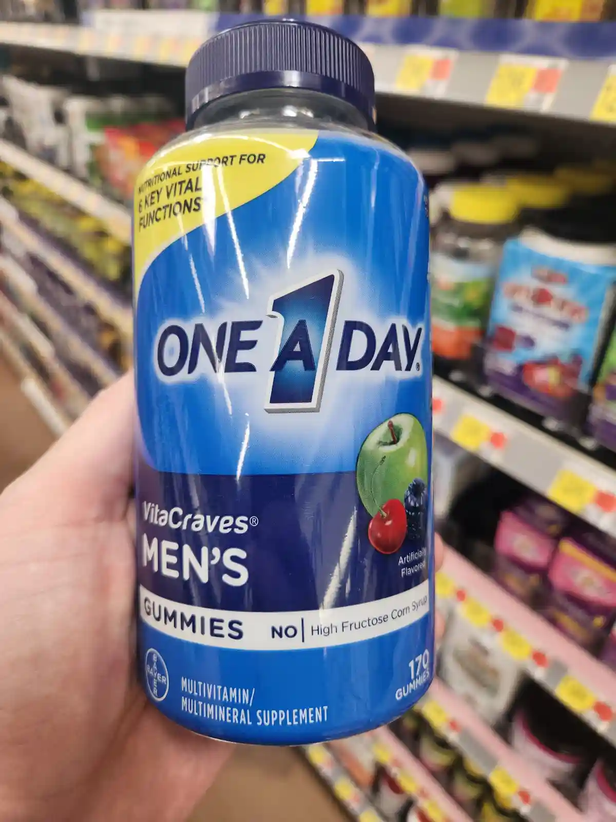 A hand holds a blue bottle of One A Day Men's Gummies in a store aisle, with the label clearly showing 'VitaCraves MEN'S…