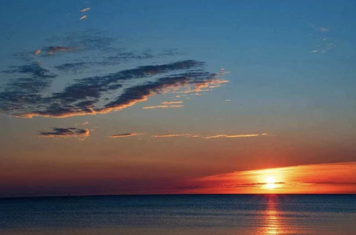 A serene sunset over the ocean with the sun low on the horizon, casting orange and red hues across the sky and water, with…