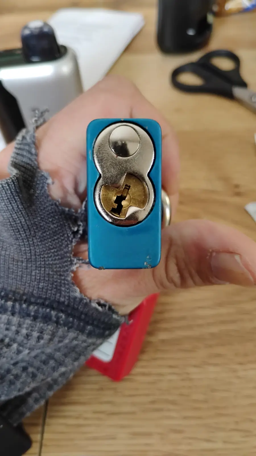 A hand holds a blue plastic lock with its internal mechanism exposed, showing a broken or damaged keyway. The background…
