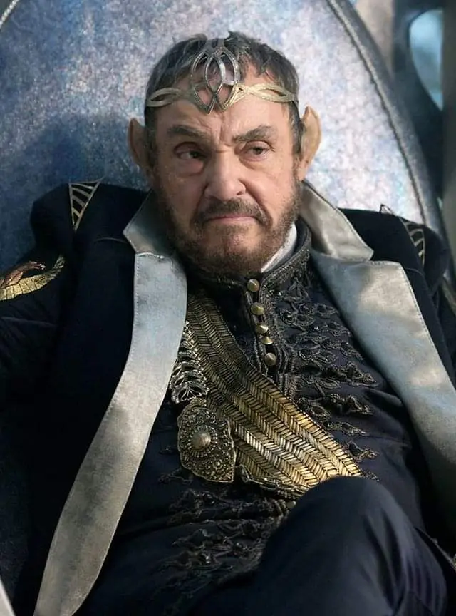 A man with pointed ears, wearing an ornate gold headband and a dark, embroidered robe with gold trim, sits in a throne-like…