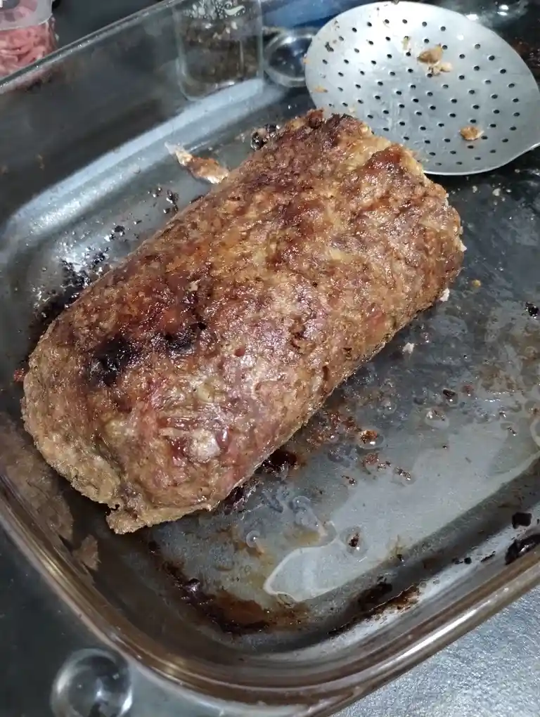 A close-up photo shows a large, browned meatloaf resting in a glass baking dish, with a metal slotted spoon nearby and some…