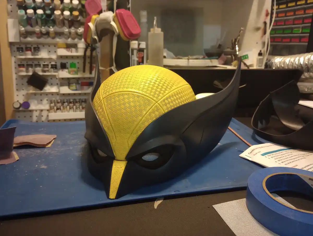 A yellow and black Wolverine-style mask sits on a blue work mat in a cluttered workshop filled with paint bottles…