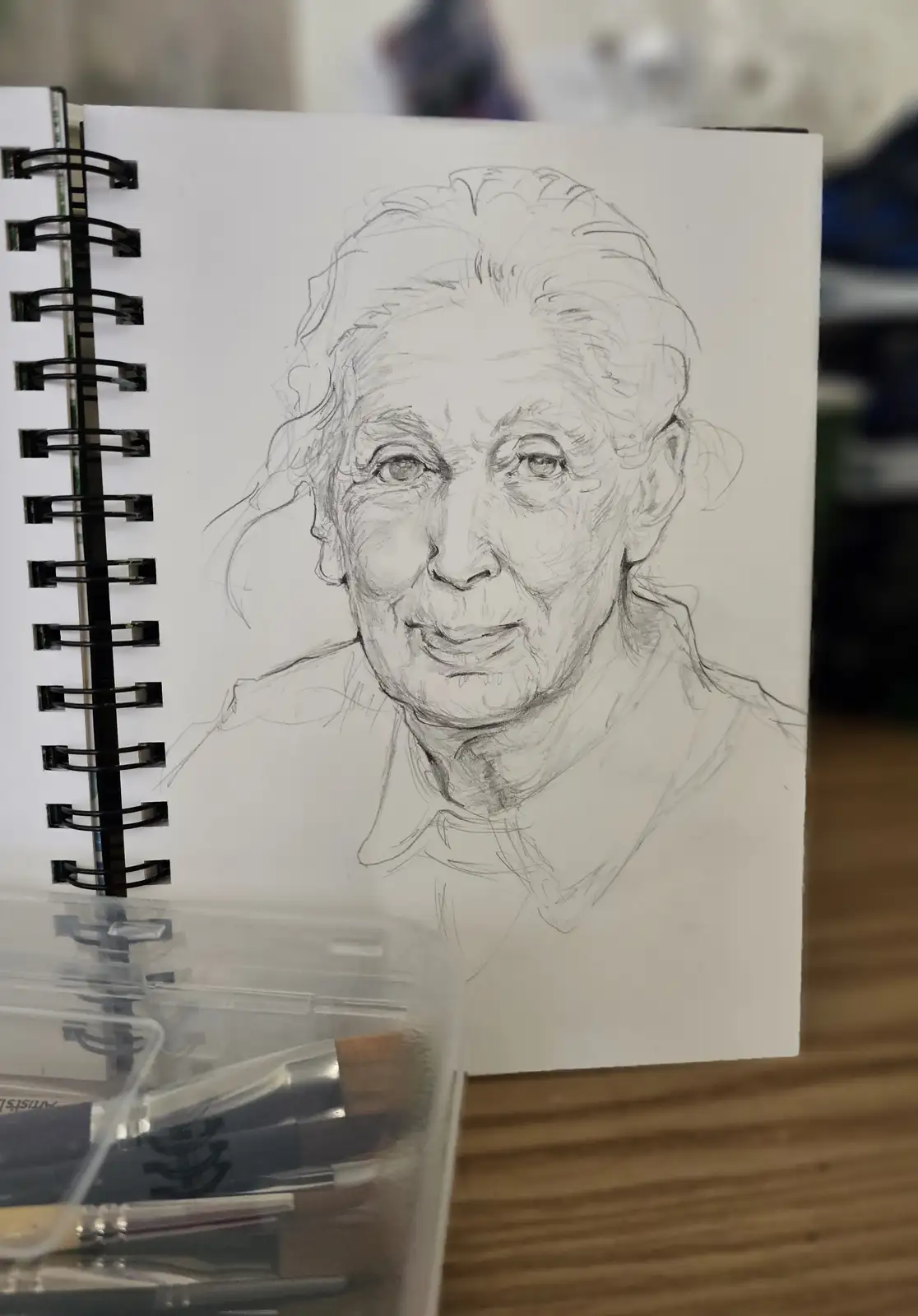 A pencil sketch of an elderly woman's face in a spiral-bound sketchbook, with a container of art brushes in the foreground…
