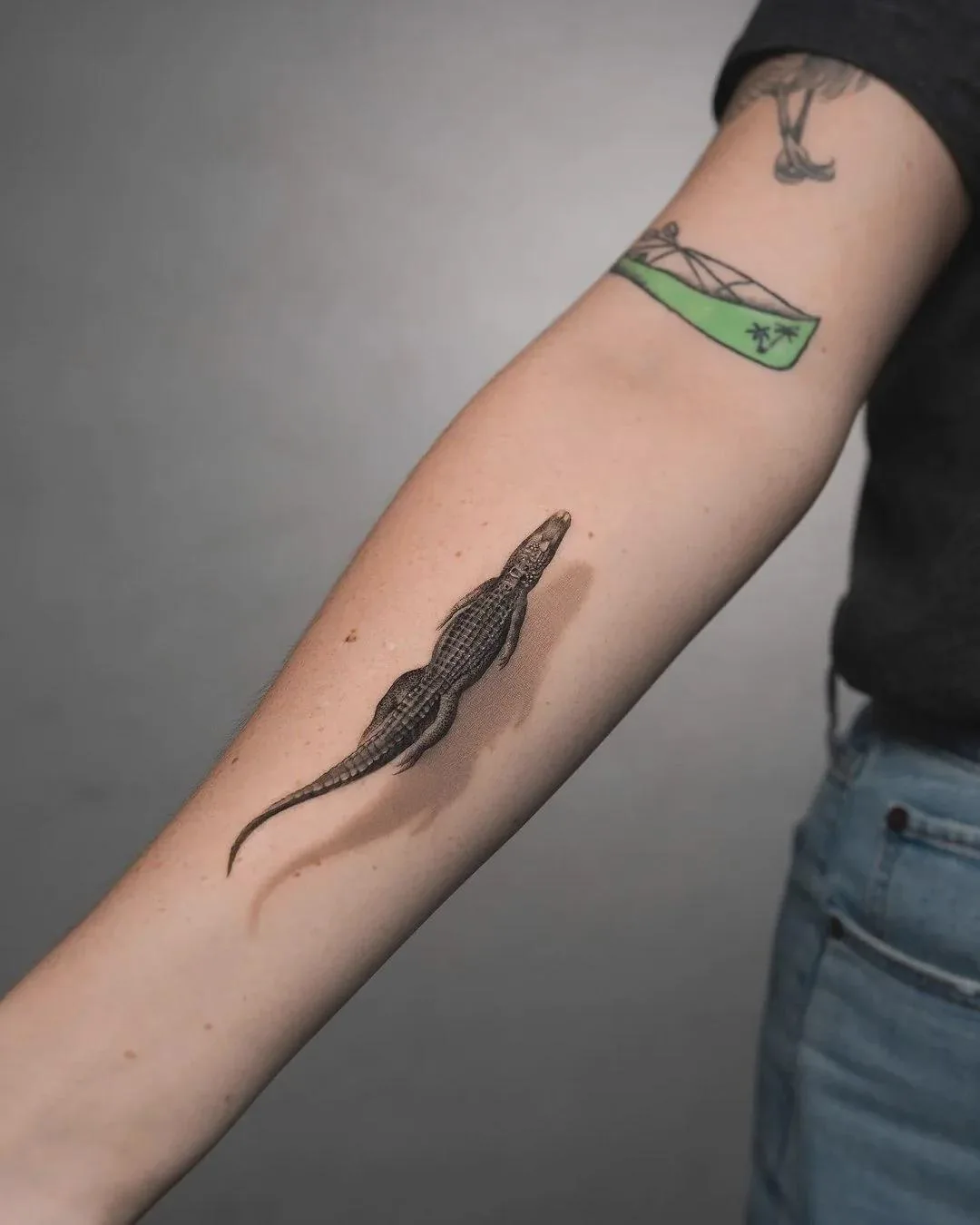 A person's forearm displays a detailed black-and-gray tattoo of a crocodile, with other tattoos visible above it. The…