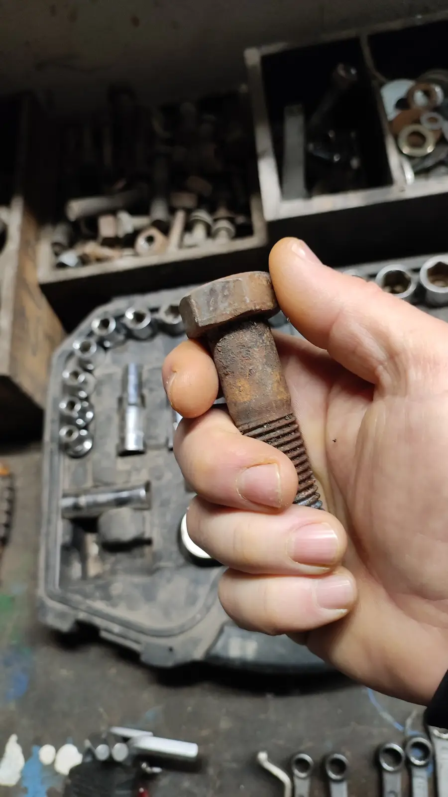 A hand holds a heavily rusted hex bolt in front of a cluttered workshop background filled with tools and hardware. The…
