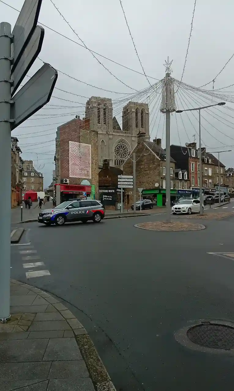A gray French police SUV is driving through a European town square with a historic church in the background, under overcast…