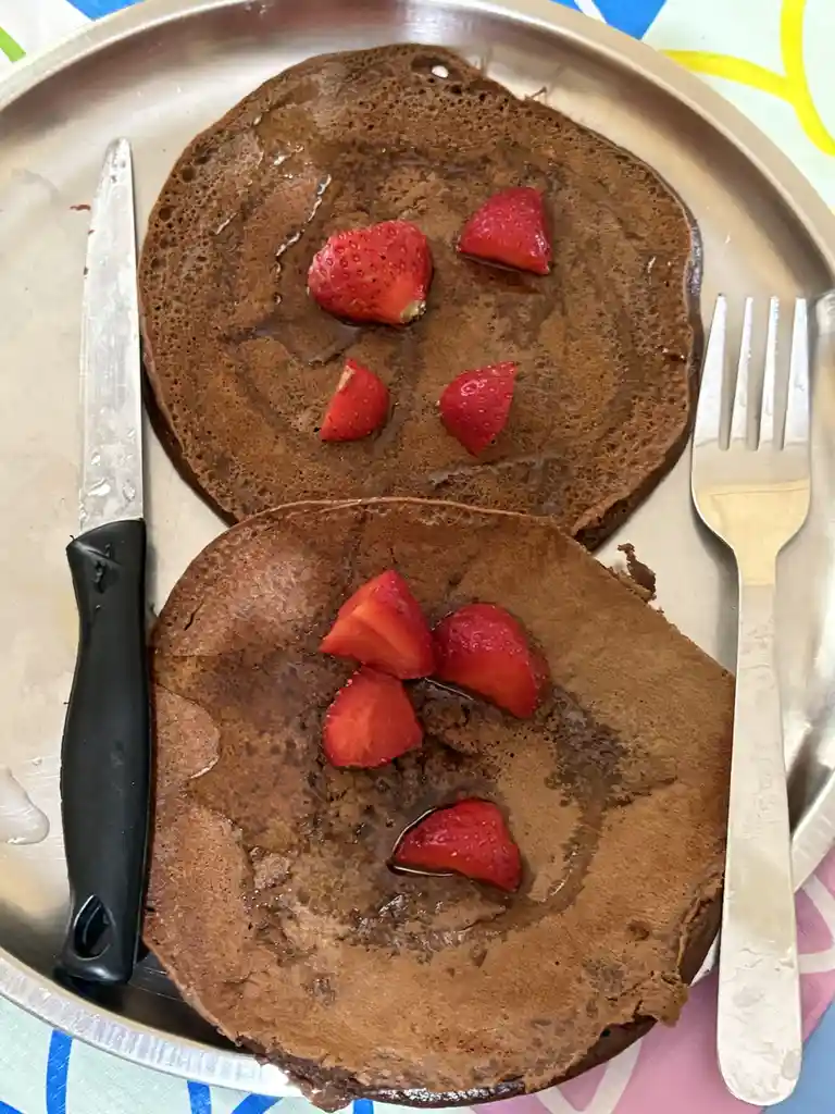Two chocolate pancakes on a metal plate are arranged to resemble crying faces, with sliced strawberries forming eyes and…