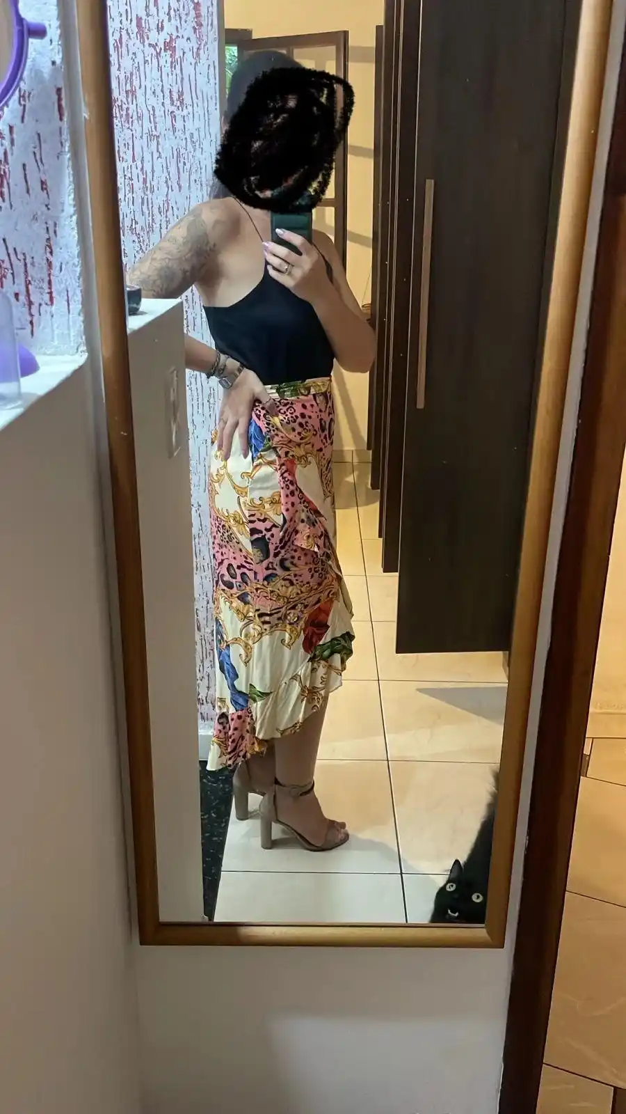 A person with a tattooed arm and obscured face takes a mirror selfie wearing a black top and a colorful, patterned skirt…