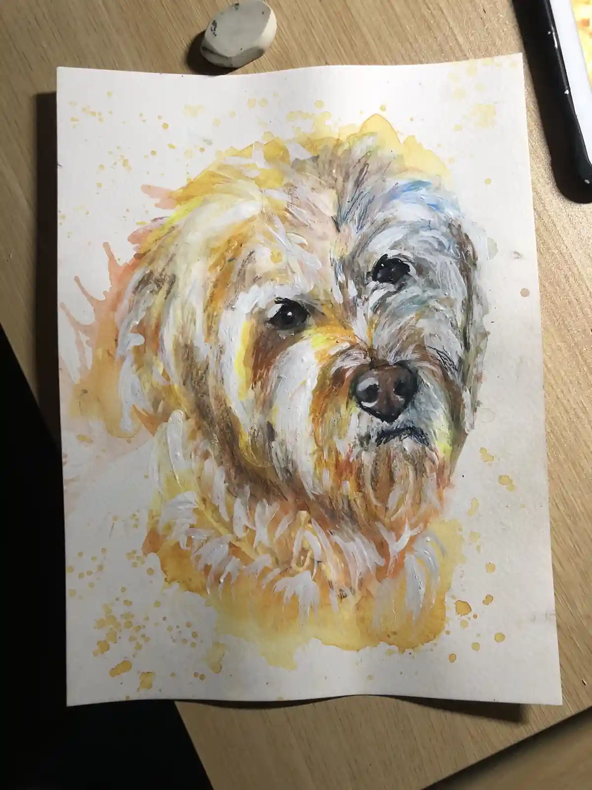 A watercolor painting of a fluffy dog's face on white paper, resting on a wooden surface with paint splatters around the…