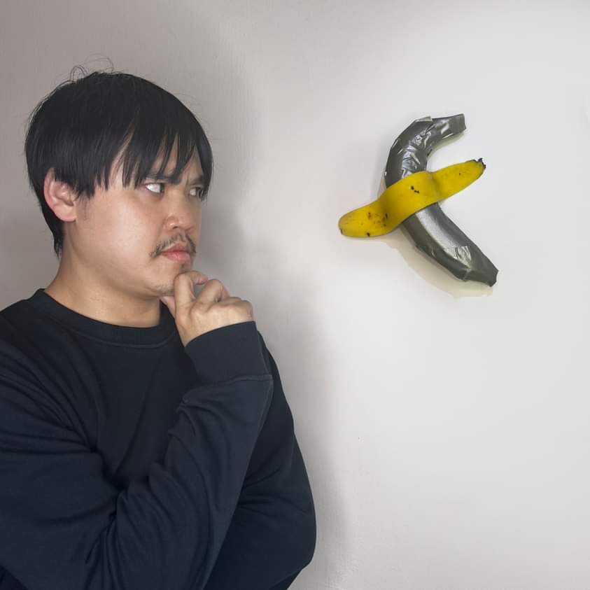 a man with dark hair and a mustache, wearing a black long-sleeved shirt, stares intently at a banana taped to a white wall…