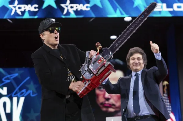 A man in a black suit and cap holds a red chainsaw guitar while standing next to Elon Musk, who is giving a thumbs-up. The…
