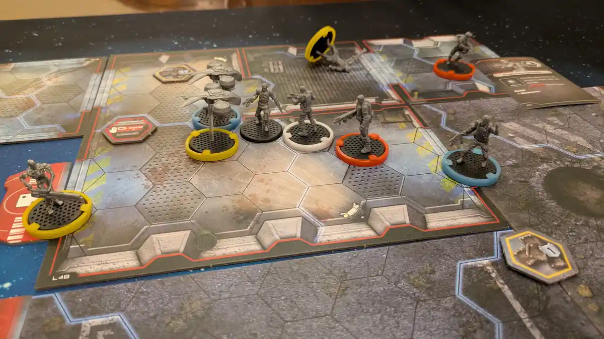 A tabletop wargame setup with grey miniatures on colored circular bases arranged on a hexagonal grid board. One miniature is…
