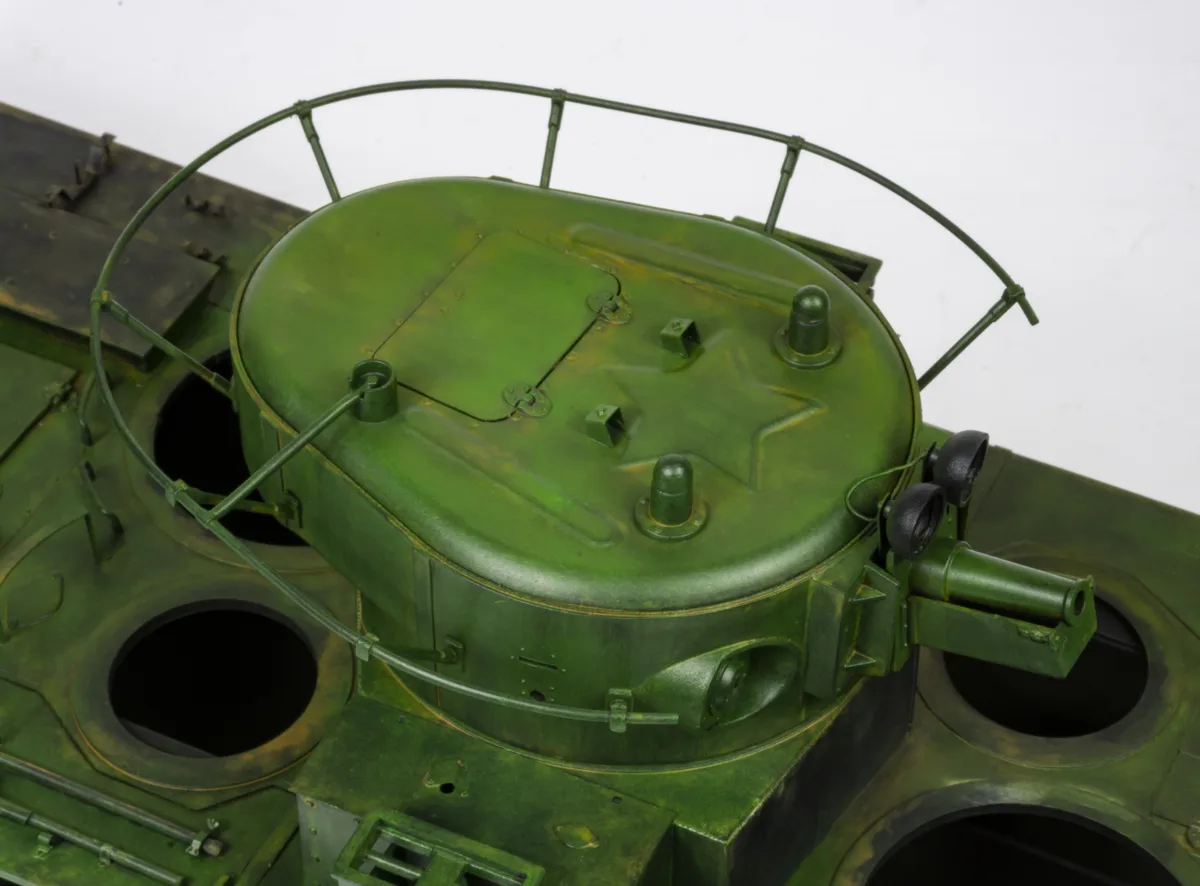 A close-up photo of a green military tank model, showing its turret with a star emblem, a wire safety railing, and a small…
