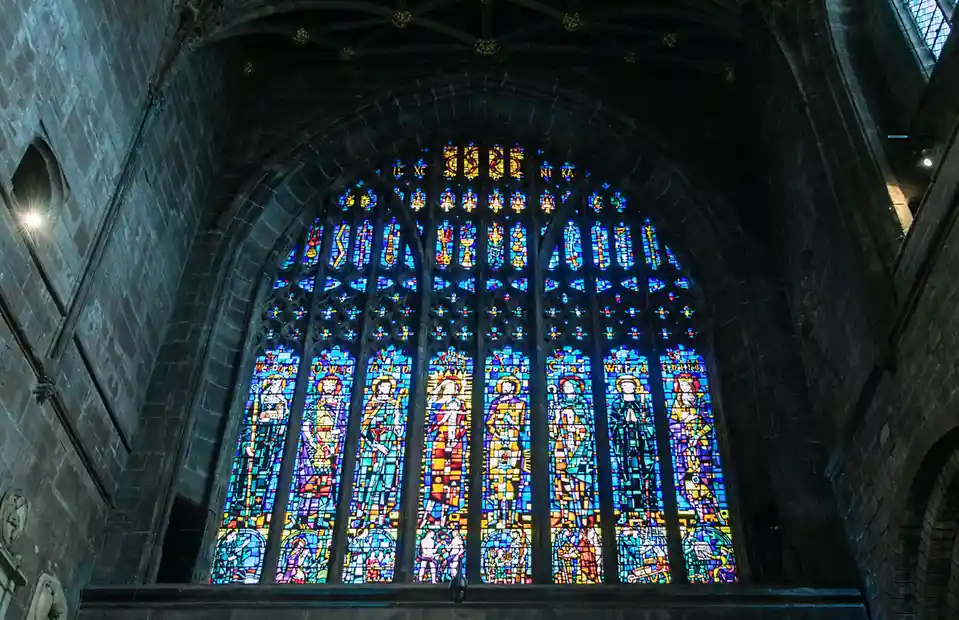 A low-angle shot inside a dark stone cathedral shows a large, ornate stained glass window with multiple panels depicting…