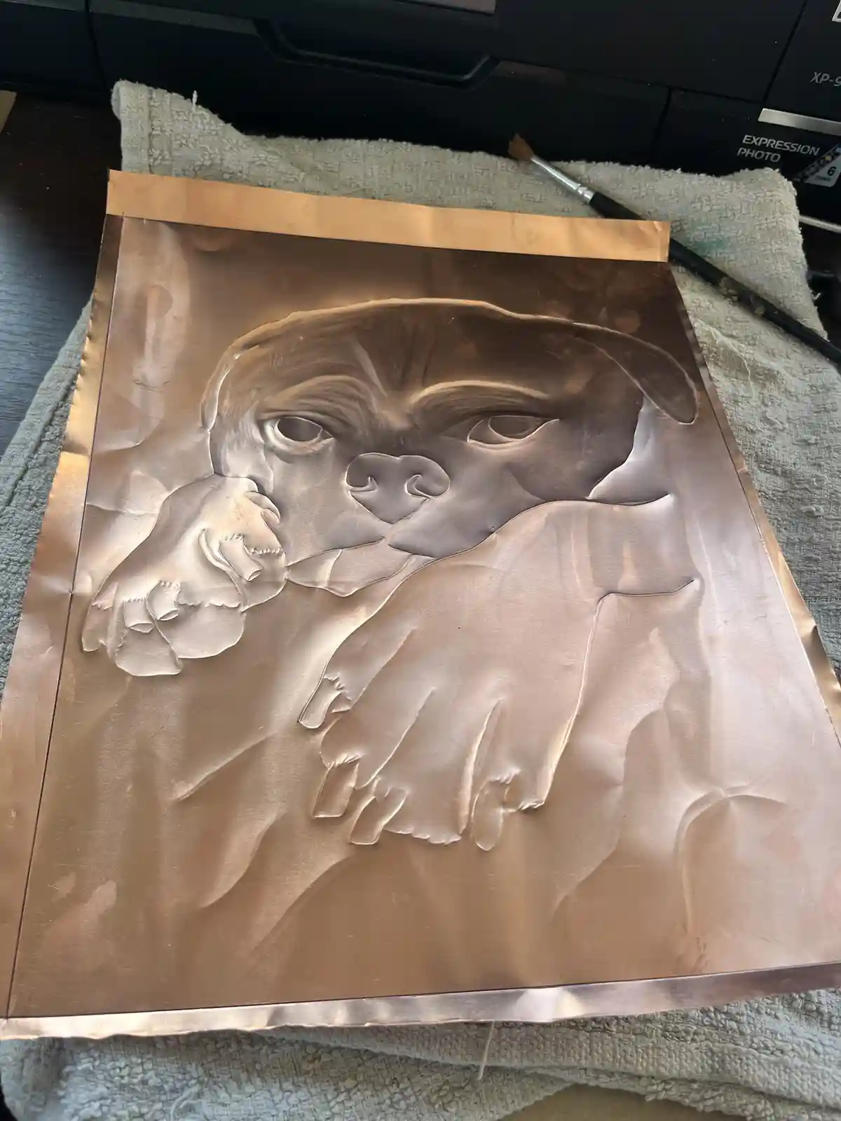 A close-up photo shows a detailed, raised relief portrait of a dog's face and paws crafted from copper sheet metal, resting…