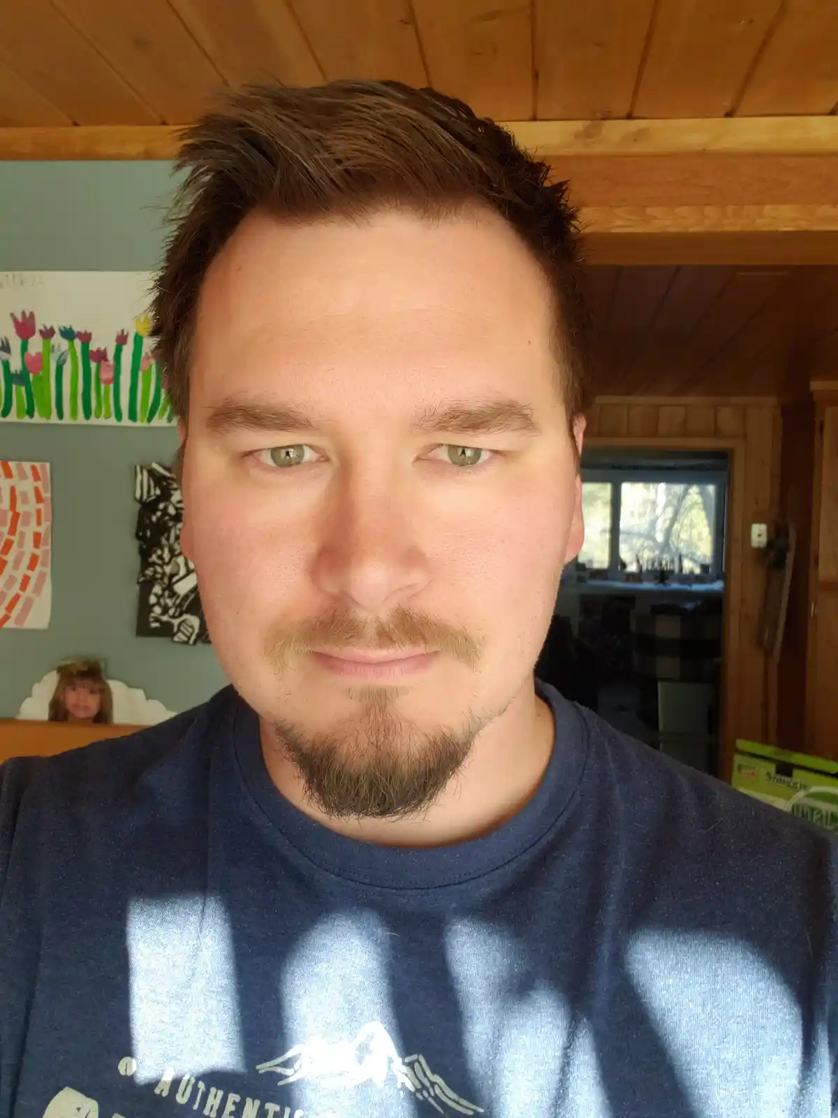 A man with a goatee and short brown hair stares directly at the camera with a neutral expression, wearing a blue t-shirt…