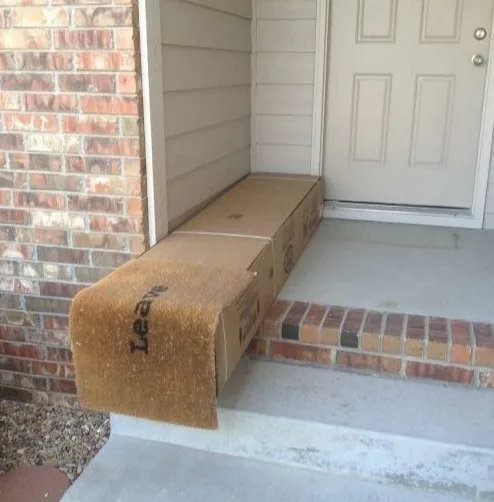 A brown doormat is visibly placed on top of a long cardboard box that is positioned on a concrete porch, directly in front…