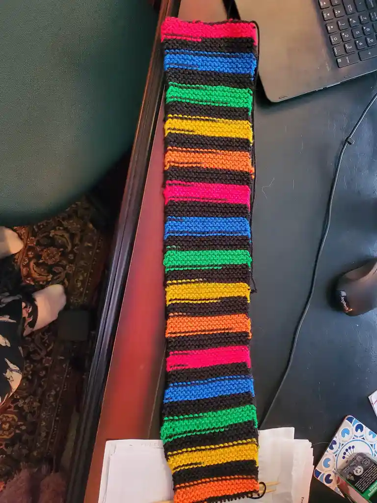 A colorful, hand-knitted scarf with repeating rainbow stripes (pink, blue, green, yellow, orange) on a black background lies…