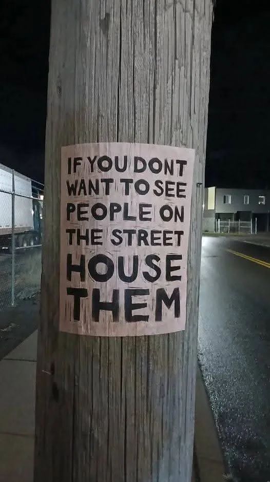 A handwritten sign taped to a wooden utility pole at night, reading 'IF YOU DONT WANT TO SEE PEOPLE ON THE STREET HOUSE…