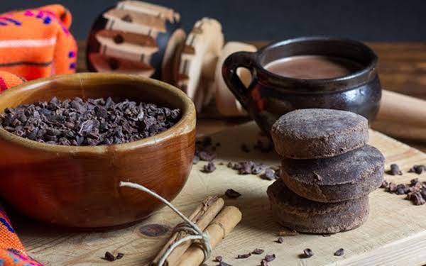 A rustic wooden board displays cocoa beans in a bowl, stacked chocolate discs, a cup of hot chocolate, and cinnamon sticks…