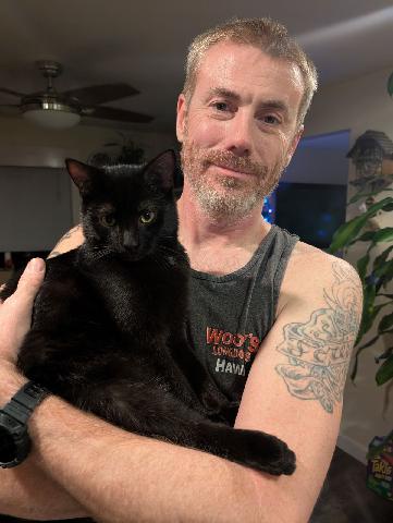 A middle-aged man with a beard and tattoos holds a black cat in his arms. He looks at the camera with a slight smirk. The…