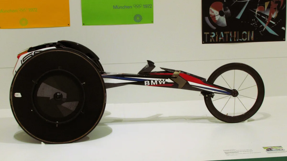 A sleek, futuristic racing wheelchair with a large front wheel and smaller rear wheel, branded with 'BMW' on its side, is…