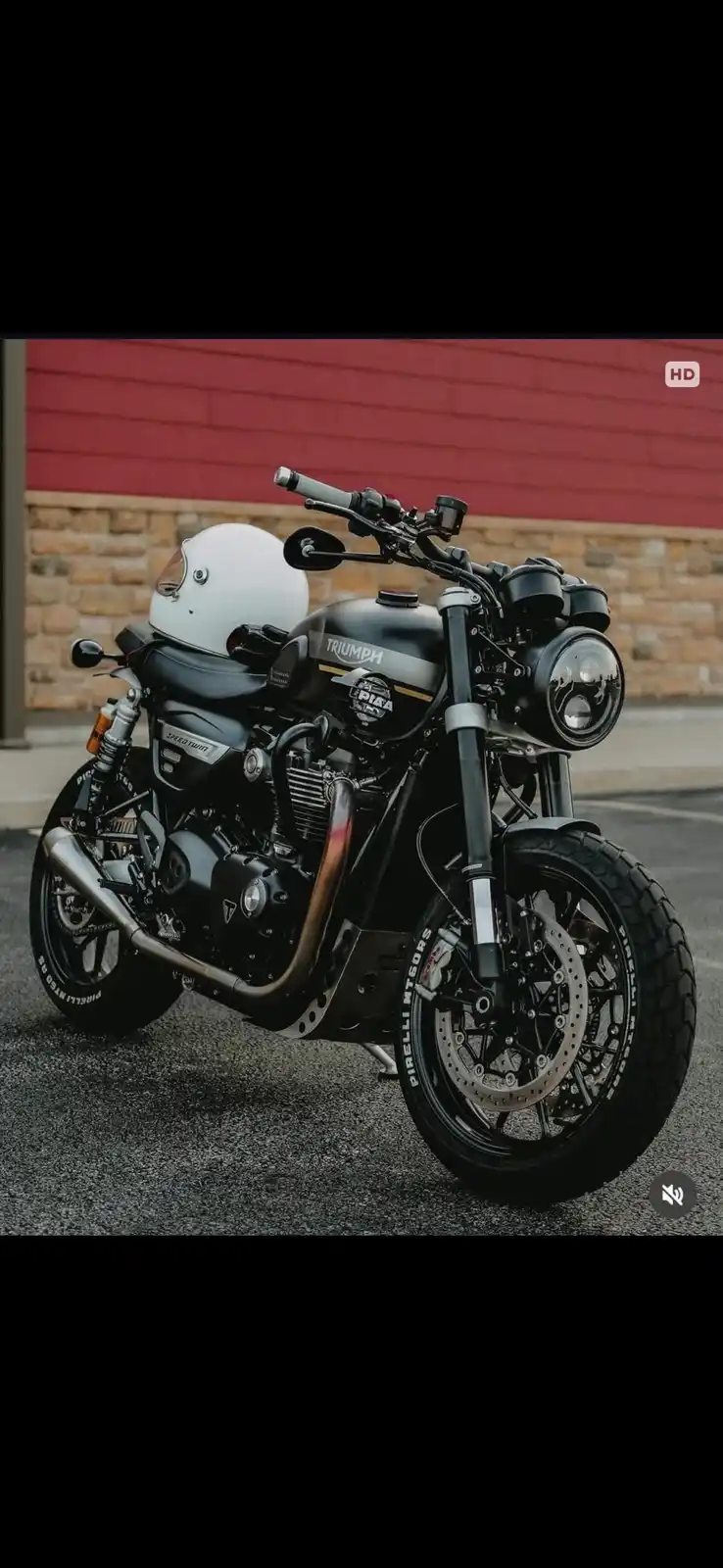 A black Triumph Speed Twin motorcycle is parked on asphalt with a white full-face helmet resting on its seat. The bike…