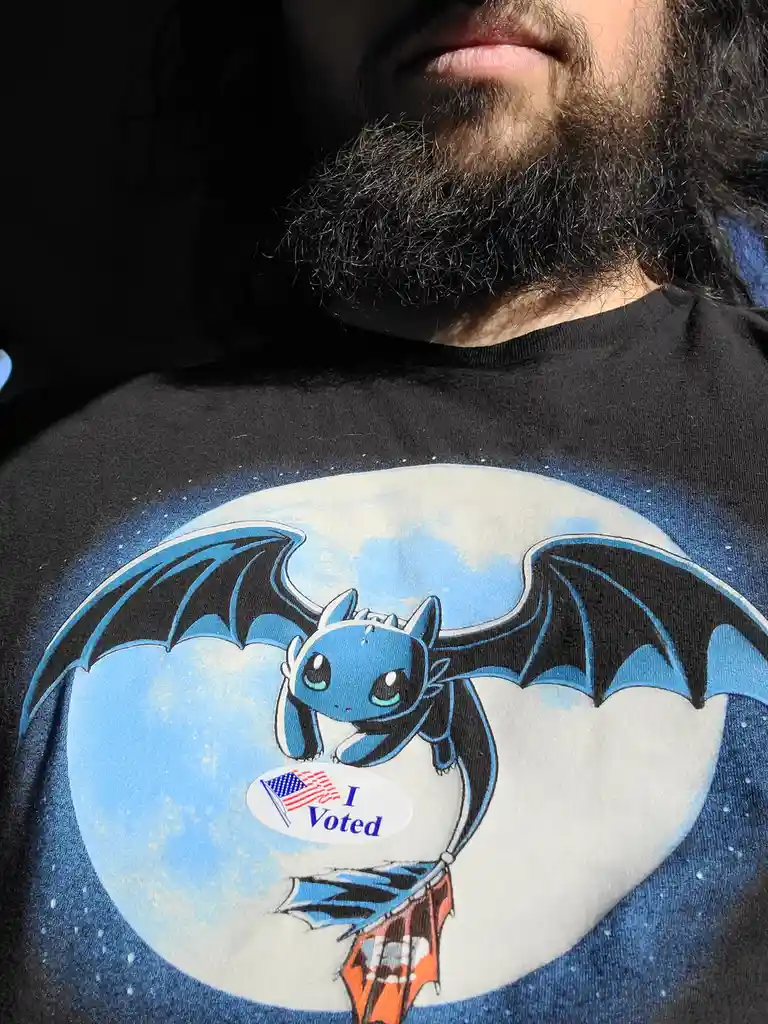 A close-up shot shows a person with a dark beard wearing a black t-shirt featuring a cartoon blue dragon with black wings…