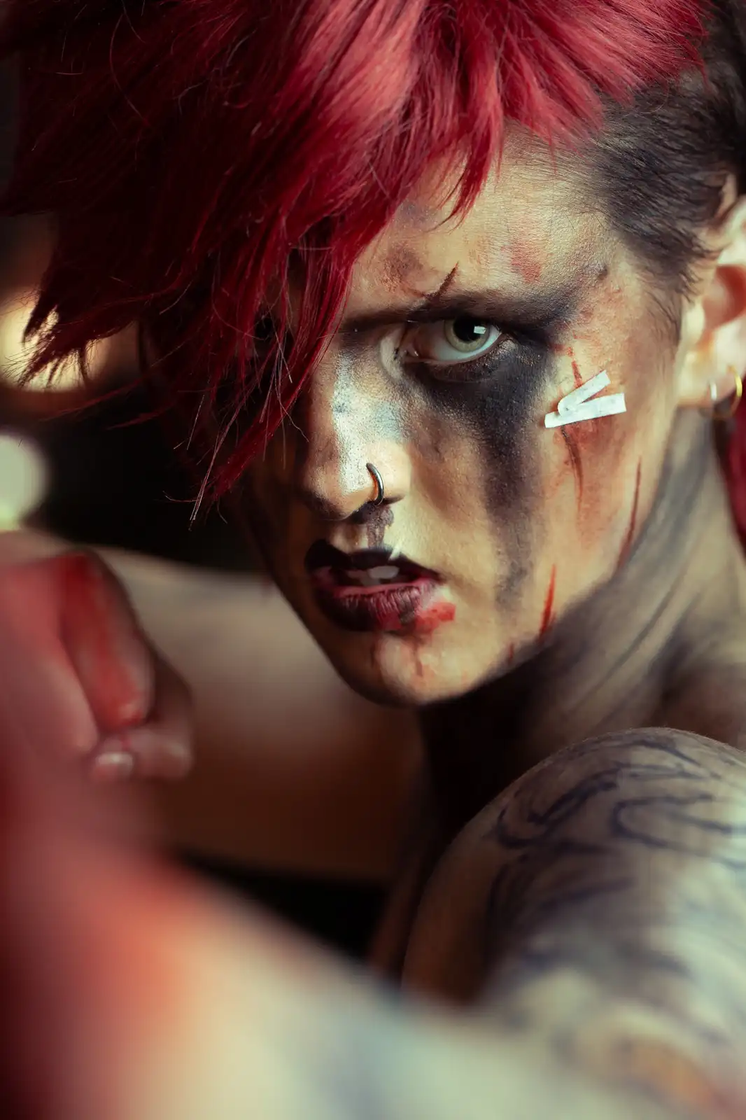 A person with red hair, heavy dark makeup, and fake blood streaks on their face stares intensely at the camera. They have a…
