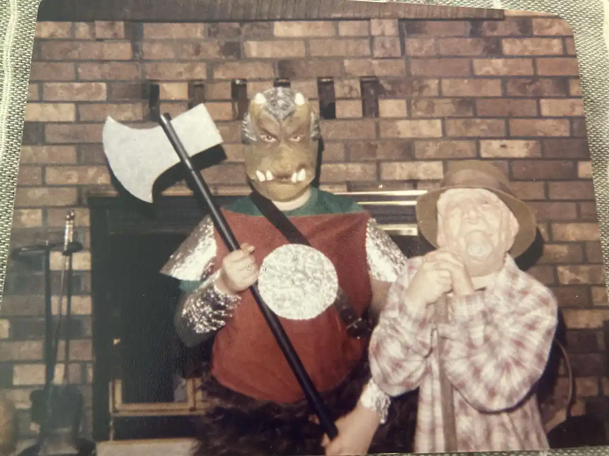 An old, grainy photo shows two people in costumes standing in front of a brick fireplace. The person on the left wears a…
