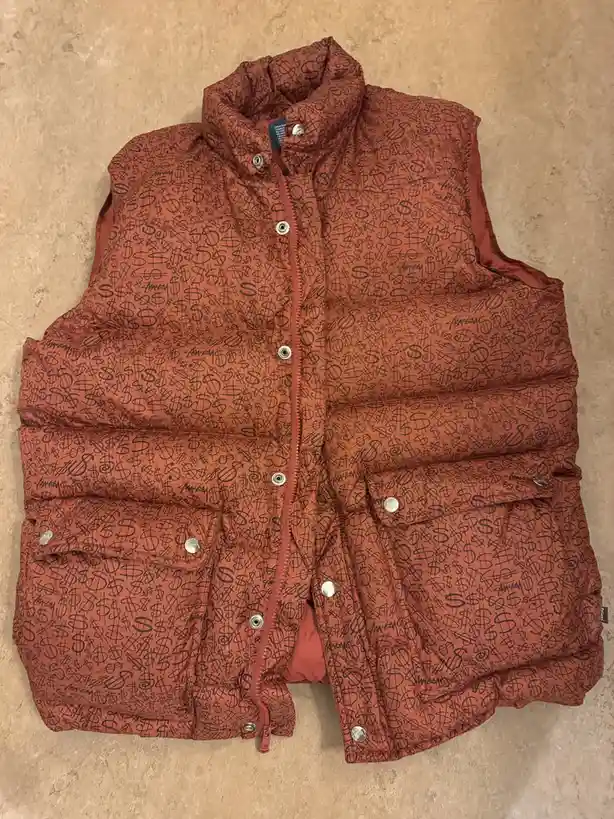 A rust-colored puffer vest with a zipper and silver snap buttons lies flat on a beige tiled floor. The entire surface of the…