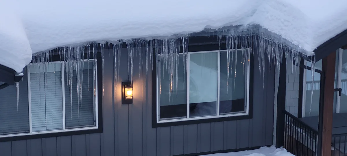 A house exterior covered in thick snow with numerous long, sharp icicles hanging from the roofline above windows. A warm…